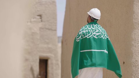 مهندس عربي خليجي سعودي مبتسم يرتدي السترة وخوذة الحماية بايماءات الفخر والاعتزاز في اليوم الوطني 23 سبتمبر، يوم بدينا 1727 م، ذكرى تأسيس الدولة السعودية الأولى 22 فبراير، رفع علم المملكة في الأماكن التراثيه الشعبيه في 11 مارس، فن العمارة القديم وبناء البيوت الطينية