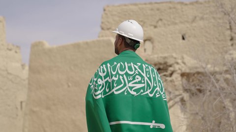 مهندس عربي خليجي سعودي مبتسم يرتدي السترة وخوذة الحماية بايماءات الفخر والاعتزاز في اليوم الوطني 23 سبتمبر، يوم بدينا 1727 م، ذكرى تأسيس الدولة السعودية الأولى 22 فبراير، رفع علم المملكة في الأماكن التراثيه الشعبيه في 11 مارس، فن العمارة القديم وبناء البيوت الطينية