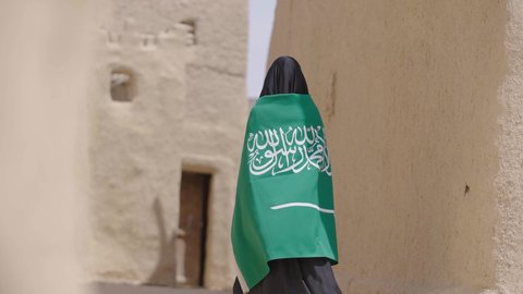 Old heritage mud houses, a symbol of national identity, a Saudi Arabian Gulf woman wearing a black abaya and burqa carrying the Kingdom's flag, celebrating the founding of the first Saudi state on February 22, the anniversary of the National Day on September 23, the day of our beginning in 1727 AD, wearing traditional folk costume