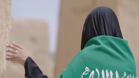 Old heritage mud houses, a symbol of national identity, a Saudi Arabian Gulf woman wearing a black abaya and burqa carrying the Kingdom's flag, celebrating the founding of the first Saudi state on February 22, the anniversary of the National Day on September 23, the day of our beginning in 1727 AD, wearing traditional folk costume