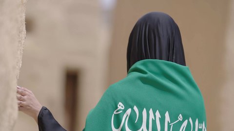 Old heritage mud houses, a symbol of national identity, a Saudi Arabian Gulf woman wearing a black abaya and burqa carrying the Kingdom's flag, celebrating the founding of the first Saudi state on February 22, the anniversary of the National Day on September 23, the day of our beginning in 1727 AD, wearing traditional folk costume