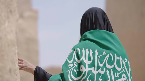 البيوت الطينية التراثية القديمة، شعار الهوية الوطنية، امرأة عربية خليجية سعودية ترتدي العباءة السوداء والبرقع تحمل علم المملكة, الاحتفال بتأسيس الدولة السعودية الأولى 22 فبراير، ذكرى اليوم الوطني في 23 سبتمبر، يوم بدينا 1727 م، ارتداء الزي الشعبي التقليدي