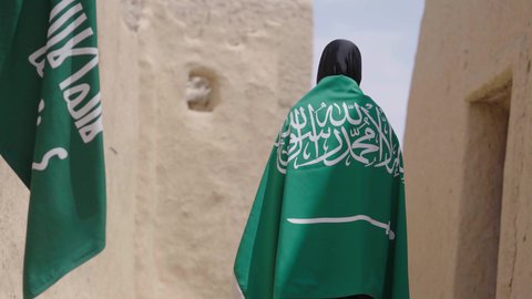 البيوت الطينية التراثية القديمة، شعار الهوية الوطنية، امرأة عربية خليجية سعودية ترتدي العباءة السوداء والبرقع تحمل علم المملكة, الاحتفال بتأسيس الدولة السعودية الأولى 22 فبراير، ذكرى اليوم الوطني في 23 سبتمبر، يوم بدينا 1727 م، ارتداء الزي الشعبي التقليدي
