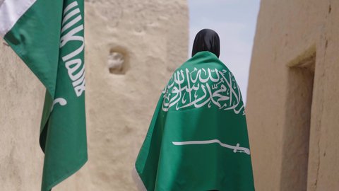 Old heritage mud houses, a symbol of national identity, a Saudi Arabian Gulf woman wearing a black abaya and burqa carrying the Kingdom's flag, celebrating the founding of the first Saudi state on February 22, the anniversary of the National Day on September 23, the day of our beginning in 1727 AD, wearing traditional folk costume