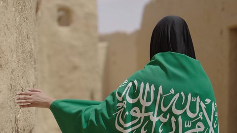 البيوت الطينية التراثية القديمة، شعار الهوية الوطنية، امرأة عربية خليجية سعودية ترتدي العباءة السوداء والبرقع تحمل علم المملكة, الاحتفال بتأسيس الدولة السعودية الأولى 22 فبراير، ذكرى اليوم الوطني في 23 سبتمبر، يوم بدينا 1727 م، ارتداء الزي الشعبي التقليدي