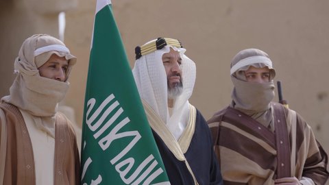 علم تأسيس المملكة العربية السعودية، إظهار الولاء والحب للوطن, مجموعة رجال عرب خليجون سعوديون يرتدون الزي الشعبي التقليدي في المناسبة الوطنية يقفون في بيت طيني قديم بإيماءات الشموخ والاعتزاز، ذكرى تأسيس الدولة السعودية الأولى 1727 م، يوم بدينا 22 فبراير