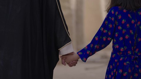A mud house in an ancient village, commemorating the founding of the first Saudi state in 1727 AD, the day of our beginning, February 22nd, a close-up of a Saudi Arabian Gulf man wearing a bisht and ghutra holding his daughter's hand as they walk around the historic neighborhood built in the old style