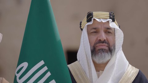 الاحتفال بيوم العلم السعودي11مارس، يوم بدينا22 فبراير, مفهوم حب الوطن والانتماء اليه, ذكرى تأسيس الدولة السعودية الأولى1727م, رجل عربي خليجي سعودي يرتدي البشت والغترة يقف في منزل طيني ويمسك علم يوم التأسيس بتعابير الشموخ، ارتداء الزي الشعبي في مناسبة وطنية