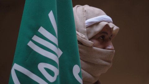 ارتداء الزي الشعبي بالمناسبة الوطنية، يوم بدينا 22 فبراير, حب الوطن والانتماء اليه, رجل عربي خليجي سعودي يرتدي البشت والغترة يقف في منزل طيني بجانب علم يوم التأسيس بتعابير الفخر والاعتزاز، الاحتفال بذكرى تأسيس الدولة السعودية الأولى 1727 م