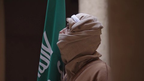 ارتداء الزي الشعبي بالمناسبة الوطنية، يوم بدينا 22 فبراير, حب الوطن والانتماء اليه, رجل عربي خليجي سعودي يرتدي البشت والغترة يقف في منزل طيني بجانب علم يوم التأسيس بتعابير الفخر والاعتزاز، الاحتفال بذكرى تأسيس الدولة السعودية الأولى 1727 م