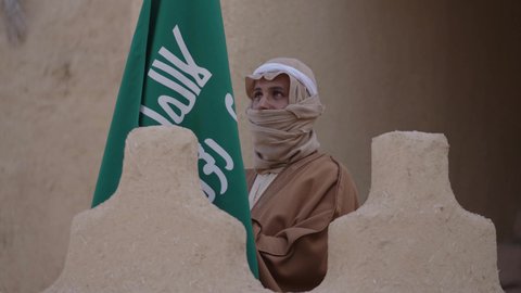 ارتداء الزي الشعبي بالمناسبة الوطنية، يوم بدينا 22 فبراير, حب الوطن والانتماء اليه, رجل عربي خليجي سعودي يرتدي البشت والغترة يقف في منزل طيني بجانب علم يوم التأسيس بتعابير الفخر والاعتزاز، الاحتفال بذكرى تأسيس الدولة السعودية الأولى 1727 م