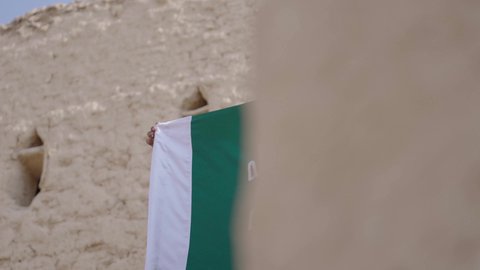 Wearing traditional costume on the national occasion, the day we started, February 22, love of the homeland and belonging to it, a Saudi Gulf Arab man wearing a bisht and ghutra stands in a mud house next to the Founding Day flag with expressions of pride and honor, celebrating the anniversary of the founding of the first Saudi state 1727 AD