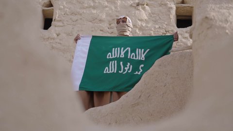 ارتداء الزي الشعبي بالمناسبة الوطنية، يوم بدينا 22 فبراير, حب الوطن والانتماء اليه, رجل عربي خليجي سعودي يرتدي البشت والغترة يقف في منزل طيني بجانب علم يوم التأسيس بتعابير الفخر والاعتزاز، الاحتفال بذكرى تأسيس الدولة السعودية الأولى 1727 م