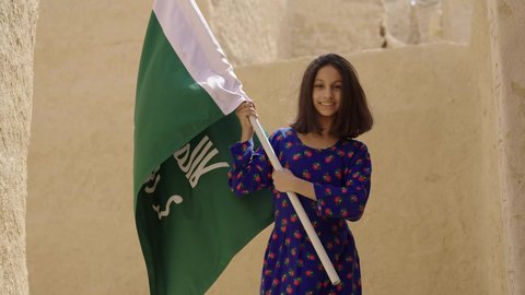 يوم العلم 11 مارس، ذكرى تأسيس الدولة السعودية الاولى 22 فبراير، فتاة عربيه خليجية سعودية مبتسمة ترتدي الزي التقليدي الشعبي تمسك بيدها العلم تتجول في منزل طيني، اليوم الوطني السعودي 23 سبتمبر، المحافظة على التراث السعودي، تربيه الأبناء على حب الوطن