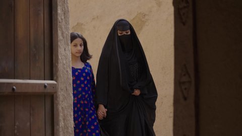 Raising children to love their country, the day we began 1727 AD, the anniversary of the founding of the first Saudi state, February 22, a Saudi Gulf Arab woman wearing a burqa and abaya walking with her daughter in a popular mud neighborhood, preserving Saudi heritage, exchanging conversations with gestures of happiness