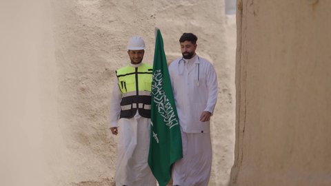 يوم بدينا 1727 م، مهن ووظائف سعودية طبية وهندسية، النهضة بالبلاد على يد ابنائها، طبيب عربي خليجي سعودي يرتدي المعطف الطبي والسماعات يحمل بيده علم المملكة يتجول برفقة مهندس في بيت طيني، إحياء ذكرى اليوم الوطني السعودي 23سبتمبر، الإحتفال بيوم العلم 11 مارس