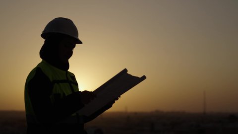 معاينة ارض المشروع, مشاريع إعمارية سعودية وطنية, تصوير من الجانب لمهندسة عربية سعودية خليجية محجبة ترتدي الخوذة وسترة الحماية تقوم بالاطلاع على المخططات, بناء الوطن بسواعد أبنائه, خلفية غروب الشمس