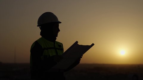 Project site inspection, Saudi national construction projects, side view of a veiled Saudi Arabian Gulf engineer wearing a helmet and a protective vest looking at the plans, building the nation with the help of its sons, sunset background