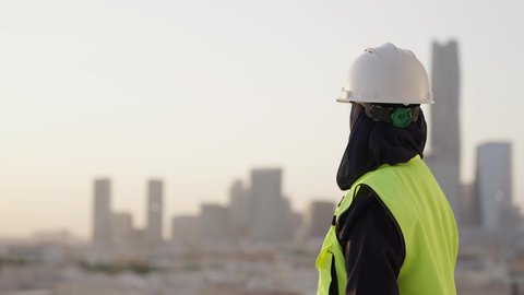 Pride and honor in the Kingdom's urban renaissance, Saudi national construction projects, close-up from the back of a veiled Saudi Arabian Gulf engineer wearing a helmet and a protective vest standing proudly, building the nation with the hands of its sons and engineers, the distinguished urban development of the capital, background of the Riyadh towers