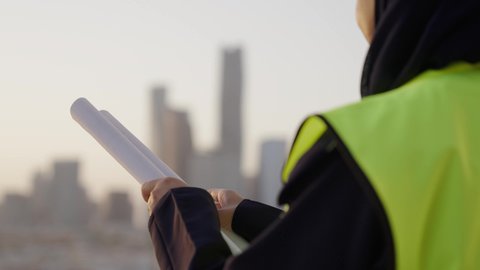 The capital's distinguished urban development, pride and honor in the Kingdom's urban renaissance, Saudi national construction projects, a close-up shot from the back of a veiled Saudi Arabian Gulf engineer wearing a helmet and a protective vest, examining the plans, building the nation with the hands of its sons, the background of the Riyadh towers