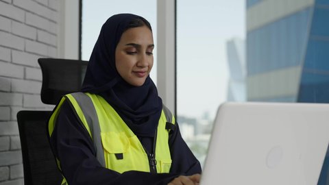 Achieving company goals, a veiled Saudi Arabian Gulf engineer wearing a protective vest works on her laptop with gestures of integration and focus, using modern technology, completing daily tasks in the office, Saudi women's professions and jobs.