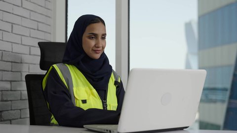 Achieving company goals, a veiled Saudi Arabian Gulf engineer wearing a protective vest works on her laptop with gestures of integration and focus, using modern technology, completing daily tasks in the office, Saudi women's professions and jobs.