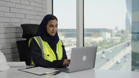 استخدام الوسائل التقنية الحديثة، تحقيق أهداف الشركة، مهندسة عربية سعودية خليجية محجبة ترتدي سترة الحماية تقوم بتدوين الملاحظات والمعلومات، العمل على جهاز الحاسوب المحمول بإيماءات الاندماج والتركيز،  إنجاز المهام اليومية في المكتب, مهن ووظائف نسائية سعودية.