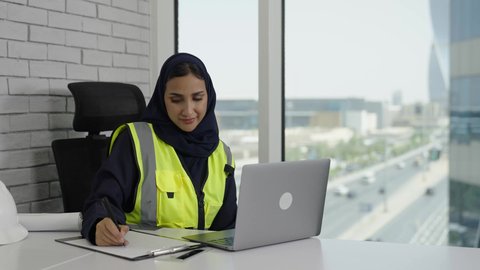 Using modern technology to achieve company goals, a veiled Saudi Arabian Gulf engineer wearing a protective vest taking notes and information, working on a laptop with gestures of integration and concentration, completing daily tasks in the office, Saudi women's professions and jobs.