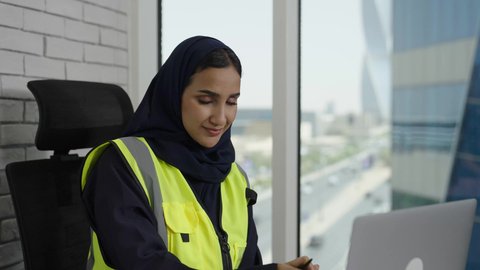 Completing daily office tasks, using modern technology, achieving company goals, close-up of a veiled Saudi Arabian Gulf female engineer wearing a protective vest taking notes and information, working on a laptop, Saudi women's professions and jobs.