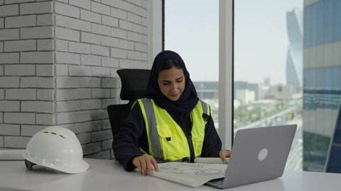 Following up and auditing engineering project strategies, the concept of engineering and architectural construction, a veiled Saudi Arabian Gulf female engineer wearing a work jacket sitting behind a desk holding an engineering plan, women's office jobs and professions, using a laptop computer