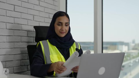 Following up and auditing engineering project strategies, the concept of engineering and architectural construction, a veiled Saudi Arabian Gulf female engineer wearing a work jacket sitting behind a desk holding an engineering plan, women's office jobs and professions, using a laptop computer