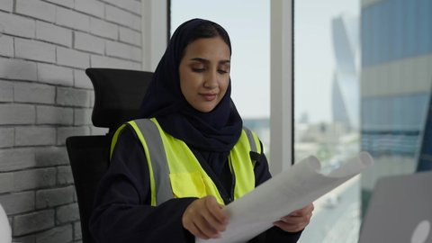 The concept of engineering and architectural construction, following up on engineering project strategies and reviewing them, a close-up of a veiled Saudi Arabian Gulf female engineer wearing a work jacket sitting behind a desk holding an engineering plan, women's office jobs and professions, using a laptop computer
