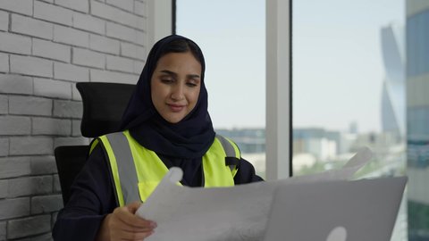 The concept of engineering and architectural construction, following up on engineering project strategies and reviewing them, a close-up of a veiled Saudi Arabian Gulf female engineer wearing a work jacket sitting behind a desk holding an engineering plan, women's office jobs and professions, using a laptop computer