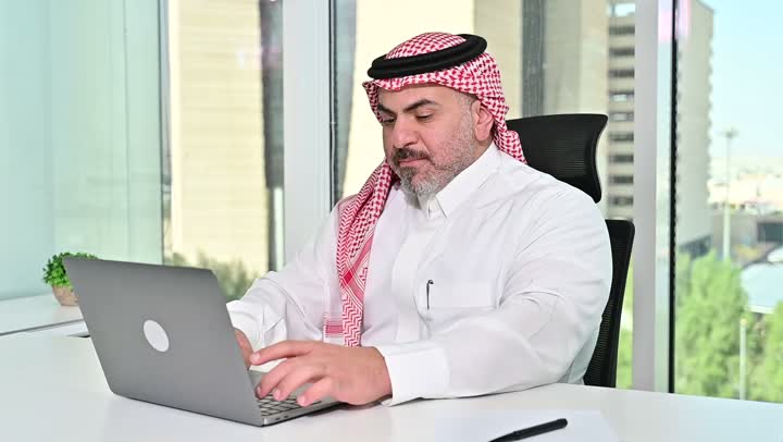 ايماءات التركيز والاندماج، مهنة ووظيفة مكتبية سعودية، كتابة وتدوين البيانات والنتائج، ادارة وانجاز المهام باستخدام جهاز تقني حديث ومتطور, تصوير مقرب لرجل عربي خليجي سعودي يرتدي الشماغ والثوب التقليدي يعمل على الحاسوب المحمول