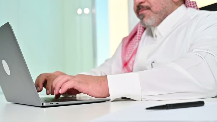 ايماءات التركيز والاندماج، مهنة ووظيفة مكتبية سعودية، كتابة وتدوين البيانات والنتائج، ادارة وانجاز المهام باستخدام جهاز تقني حديث ومتطور, تصوير مقرب لرجل عربي خليجي سعودي يرتدي الشماغ والثوب التقليدي يعمل على الحاسوب المحمول