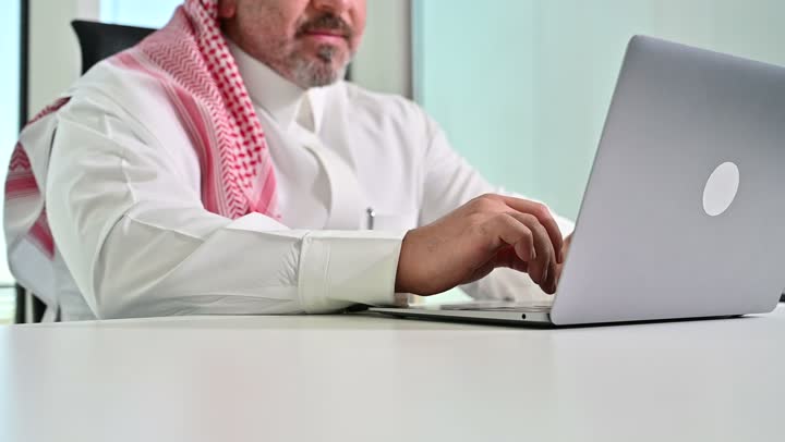 ايماءات التركيز والاندماج، مهنة ووظيفة مكتبية سعودية، كتابة وتدوين البيانات والنتائج، ادارة وانجاز المهام باستخدام جهاز تقني حديث ومتطور, تصوير مقرب لرجل عربي خليجي سعودي يرتدي الشماغ والثوب التقليدي يعمل على الحاسوب المحمول