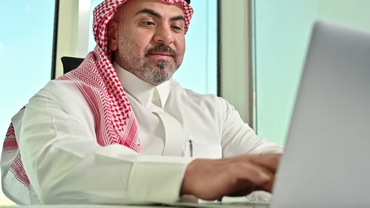 Gestures of concentration and integration, Saudi office profession and job, writing and recording data and results, managing and completing tasks using a modern and advanced technical device, close-up of a Saudi Arabian Gulf man wearing a traditional shemagh and thobe working on a laptop