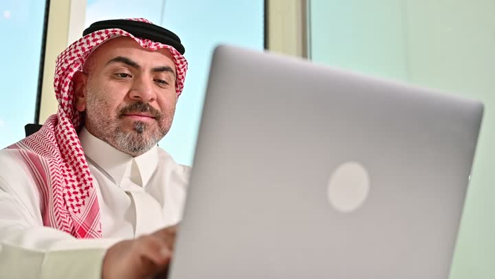 ايماءات التركيز والاندماج، مهنة ووظيفة مكتبية سعودية، كتابة وتدوين البيانات والنتائج، ادارة وانجاز المهام باستخدام جهاز تقني حديث ومتطور, تصوير مقرب لرجل عربي خليجي سعودي يرتدي الشماغ والثوب التقليدي يعمل على الحاسوب المحمول