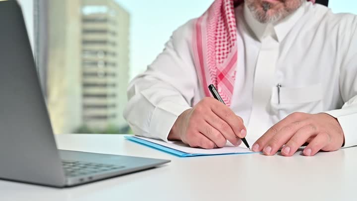Saudi office profession and job, writing and recording data and results, managing and completing tasks using a modern and advanced technical device, close-up of a Saudi Arabian Gulf man wearing a traditional shemagh and thobe working on a laptop, gestures of concentration and integration