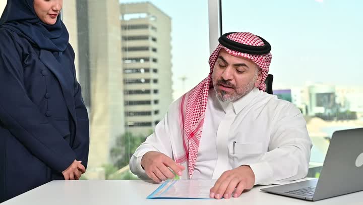 العمل على الحاسوب المحمول، مهنة ووظيفة مكتبية سعودية، كتابة وتدوين البيانات والنتائج، ادارة وانجاز المهام باستخدام جهاز تقني حديث ومتطور, تصوير مقرب لرجل عربي خليجي سعودي يرتدي الشماغ والثوب التقليدي وتقف بجانبه السكرتيرة