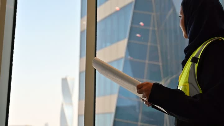 مشاريع إعمارية سعودية وطنية, تصوير مقرب لمهندسة عربية سعودية خليجية محجبة ترتدي الخوذة وسترة الحماية تقوم بالاطلاع على المخططات, بناء الوطن بسواعد أبنائه, التطور العمراني المتميز للعاصمة, الافتخار والاعتزاز بالنهضة العمرانية للمملكة, خلفية أبراج الرياض