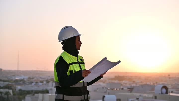 مشاريع إعمارية سعودية وطنية, تصوير مقرب لمهندسة عربية سعودية خليجية محجبة ترتدي الخوذة وسترة الحماية تقوم بالاطلاع على المخططات, بناء الوطن بسواعد أبنائه, التطور العمراني المتميز للعاصمة, الافتخار والاعتزاز بالنهضة العمرانية للمملكة, خلفية أبراج الرياض