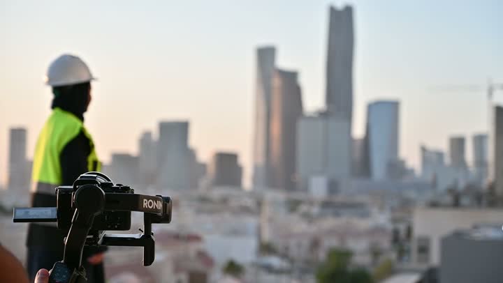 لقطات فنية فوتوغرافية باستخدام الكاميرا، مشاريع إعمارية سعودية وطنية,  مصور يقوم بتصوير مهندسة عربية سعودية خليجية محجبة ترتدي الخوذة وسترة الحماية تقوم بالاطلاع على المخططات, بناء الوطن بسواعد أبنائه, التطور العمراني المتميز للعاصمة, خلفية أبراج الرياض