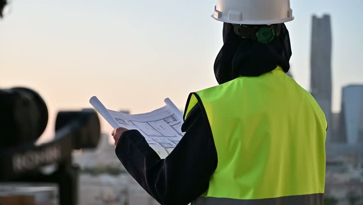 لقطات فنية فوتوغرافية باستخدام الكاميرا، مشاريع إعمارية سعودية وطنية,  مصور يقوم بتصوير مهندسة عربية سعودية خليجية محجبة ترتدي الخوذة وسترة الحماية تقوم بالاطلاع على المخططات, بناء الوطن بسواعد أبنائه, التطور العمراني المتميز للعاصمة, خلفية أبراج الرياض