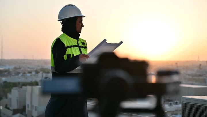 Artistic photography shots using a camera, Saudi national construction projects, a photographer photographing a veiled Saudi Arabian Gulf engineer wearing a helmet and a protective vest while examining the plans, building the nation with the hands of its sons, the distinguished urban development of the capital, the background of the Riyadh towers