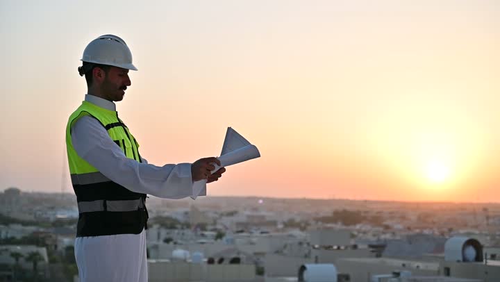 الاهتمام بالتطور العمراني بسواعد المهندسين السعوديين, مهندس عربي خليجي سعودي يرتدي سترة العمل وخوذة الحماية البيضاء يقف بفخر واعتزاز، مفهوم الهندسة والإنشاءات المعمارية، مشاريع إعمارية سعودية وطنية, خلفية الأبراج وناطحات السحاب في مدينة الرياض