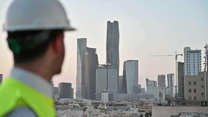 الاهتمام بالتطور العمراني بسواعد المهندسين السعوديين, مهندس عربي خليجي سعودي يرتدي سترة العمل وخوذة الحماية البيضاء يقف بفخر واعتزاز، مفهوم الهندسة والإنشاءات المعمارية، مشاريع إعمارية سعودية وطنية, خلفية الأبراج وناطحات السحاب في مدينة الرياض