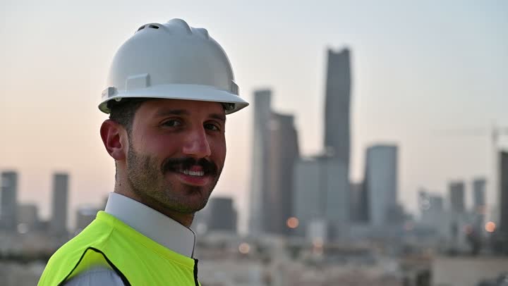 الاهتمام بالتطور العمراني بسواعد المهندسين السعوديين, مهندس عربي خليجي سعودي يرتدي سترة العمل وخوذة الحماية البيضاء يقف بفخر واعتزاز، مفهوم الهندسة والإنشاءات المعمارية، مشاريع إعمارية سعودية وطنية, خلفية الأبراج وناطحات السحاب في مدينة الرياض