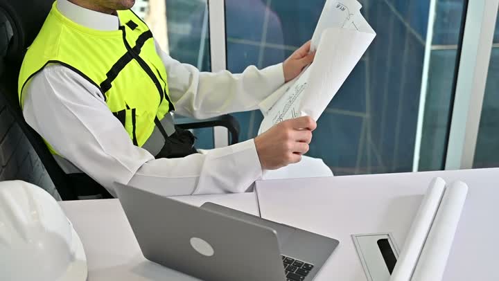Saudi national construction projects, working on a laptop, taking care of urban development with the help of Saudi engineers, a Saudi Gulf Arab engineer wearing a work jacket and a white protective helmet completing daily tasks in the office, the concept of architectural engineering and construction.