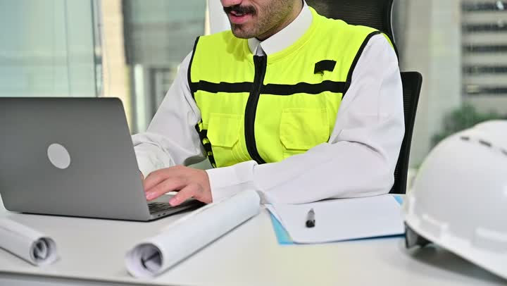 العمل على الحاسوب المحمول، الاهتمام بالتطور العمراني بسواعد المهندسين السعوديين, مهندس عربي خليجي سعودي يرتدي سترة العمل وخوذة الحماية البيضاء ينجز المهام اليومية في المكتب ، مفهوم الهندسة والإنشاءات المعمارية، مشاريع إعمارية سعودية وطنية